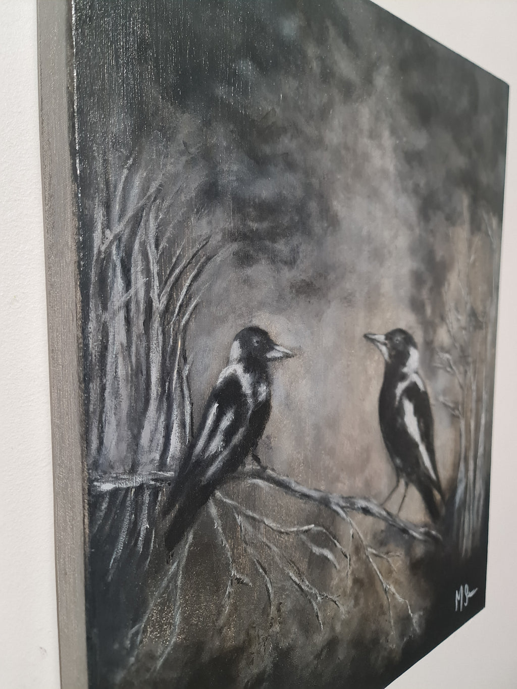 A canvas painting of two black-and-white birds perched on bare branches in a dark, smoky gray landscape, shown hanging on a light wall.