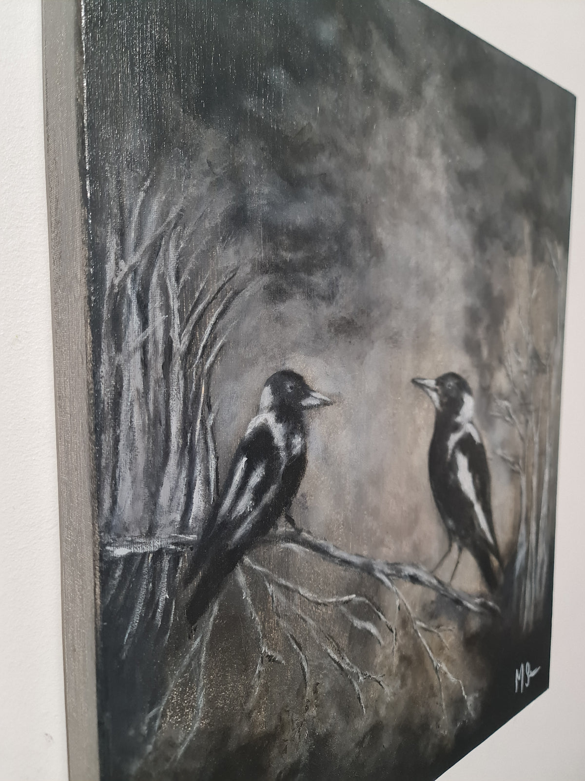 A canvas painting of two black-and-white birds perched on bare branches in a dark, smoky gray landscape, shown hanging on a light wall.